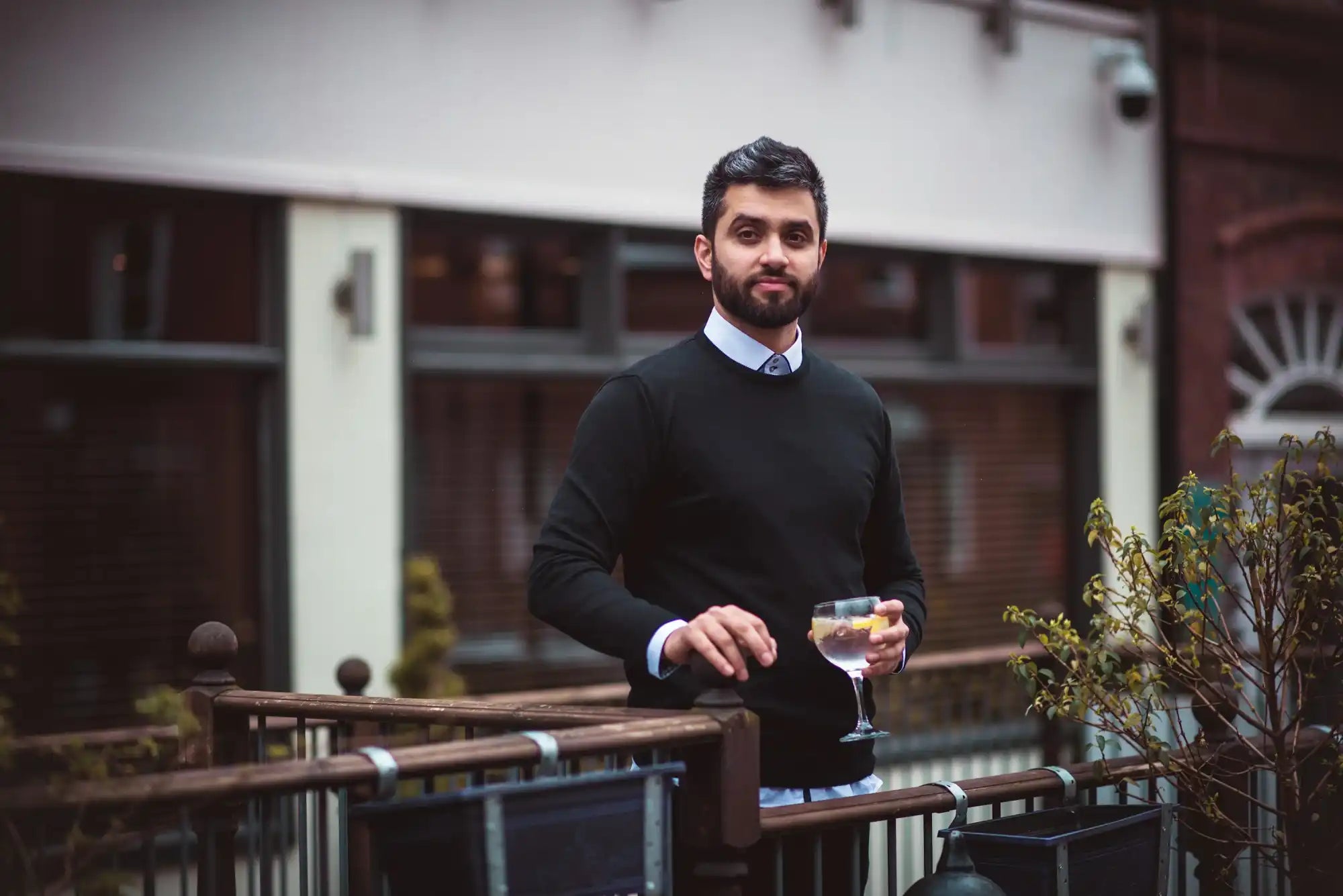 Eine gut gekleidete Person in einem schwarzen Pullover, die auf einem Balkon ein Cocktailglas hält